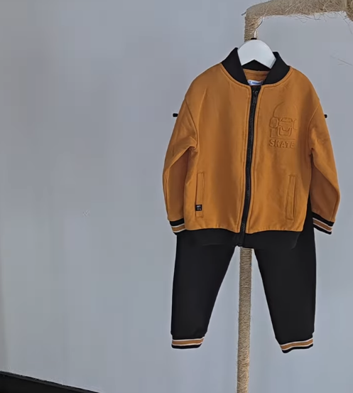 Conjunto de ropa infantil de color naranja y negro, compuesto por una chaqueta con cierre y pantalones, exhibido en una percha. La chaqueta presenta un diseño moderno con bordado de la palabra "SKATE" en el pecho. Ideal para actividades al aire libre y estilo urbano.