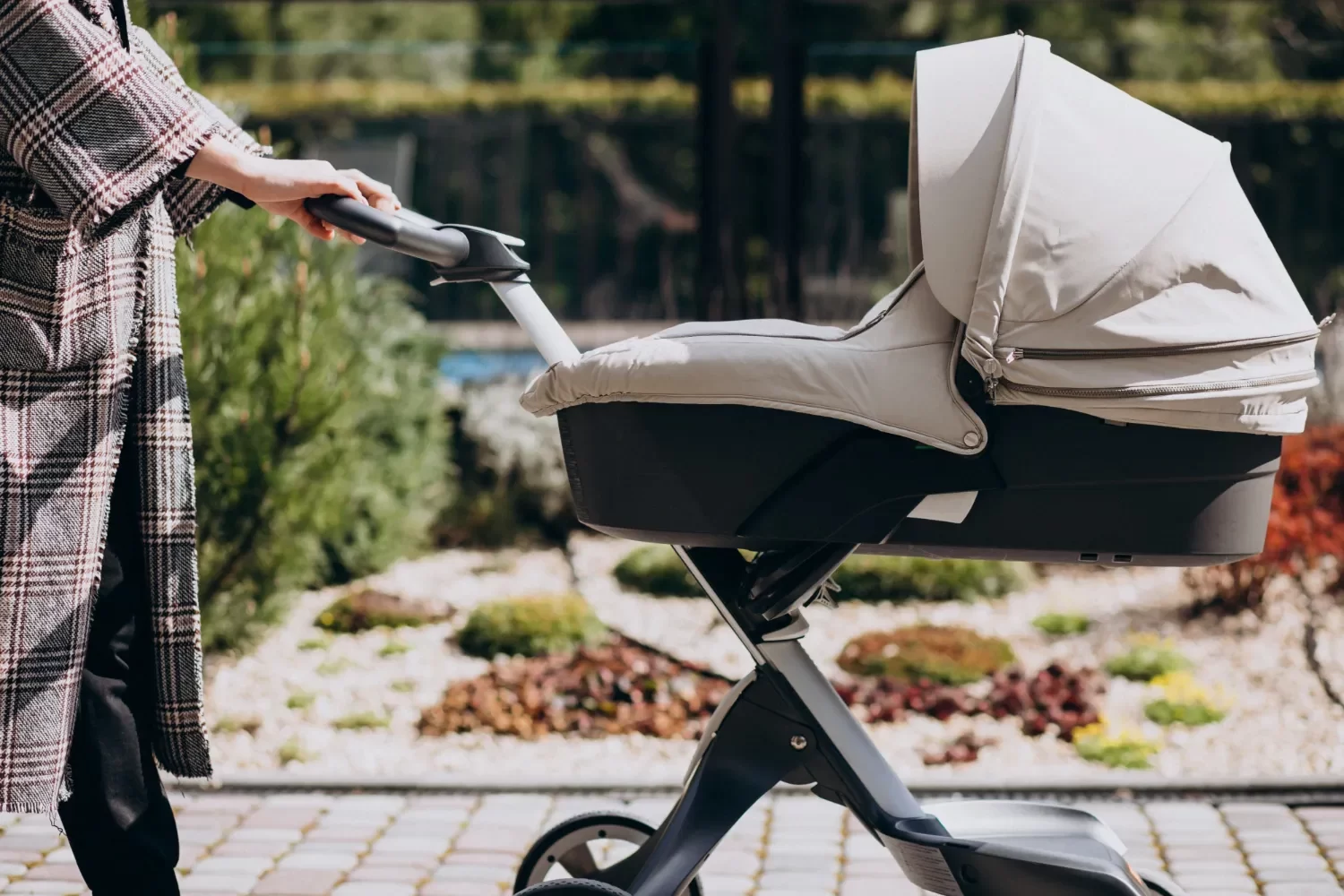 Madre empujando un cochecito de bebé elegante y moderno en un entorno ajardinado, con un diseño ergonómico y color neutro.