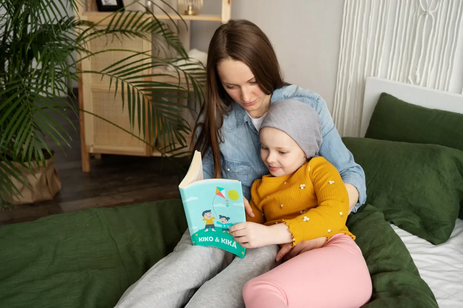Madre e hija leyendo juntas un libro titulado "Kiko & Kika" en una cama decorada con sábanas verdes. La niña, con gorro gris y ropa amarilla, muestra interés por la historia, mientras que la madre, con camisa de mezclilla, la acompaña en la lectura. El entorno es acogedor, con plantas decorativas al fondo.