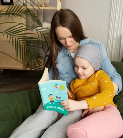 Madre e hija leyendo juntas un libro titulado "Kiko & Kika" en una cama decorada con sábanas verdes. La niña, con gorro gris y ropa amarilla, muestra interés por la historia, mientras que la madre, con camisa de mezclilla, la acompaña en la lectura. El entorno es acogedor, con plantas decorativas al fondo.