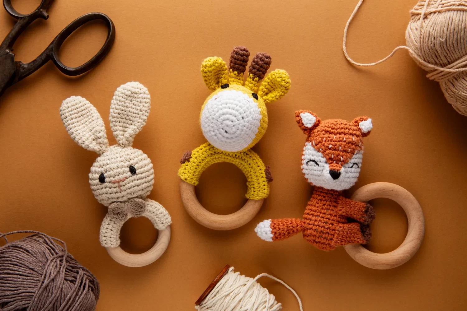 Juguetes de madera y ganchillo en forma de animales: conejo, jirafa y zorro, sobre un fondo marrón con ovillos de hilo y tijeras. Ideal para bebés y decoración.